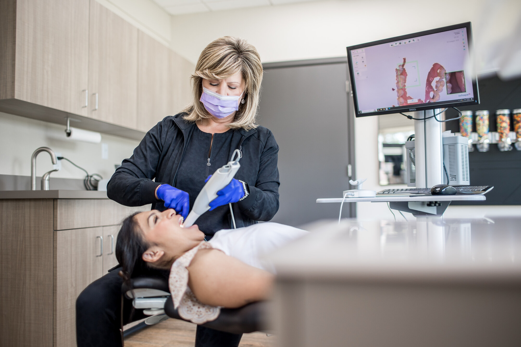 Orthodontic team member using a handheld digital scanner while a patient reclines beside a monitor displaying a three-dimensional dental scan.