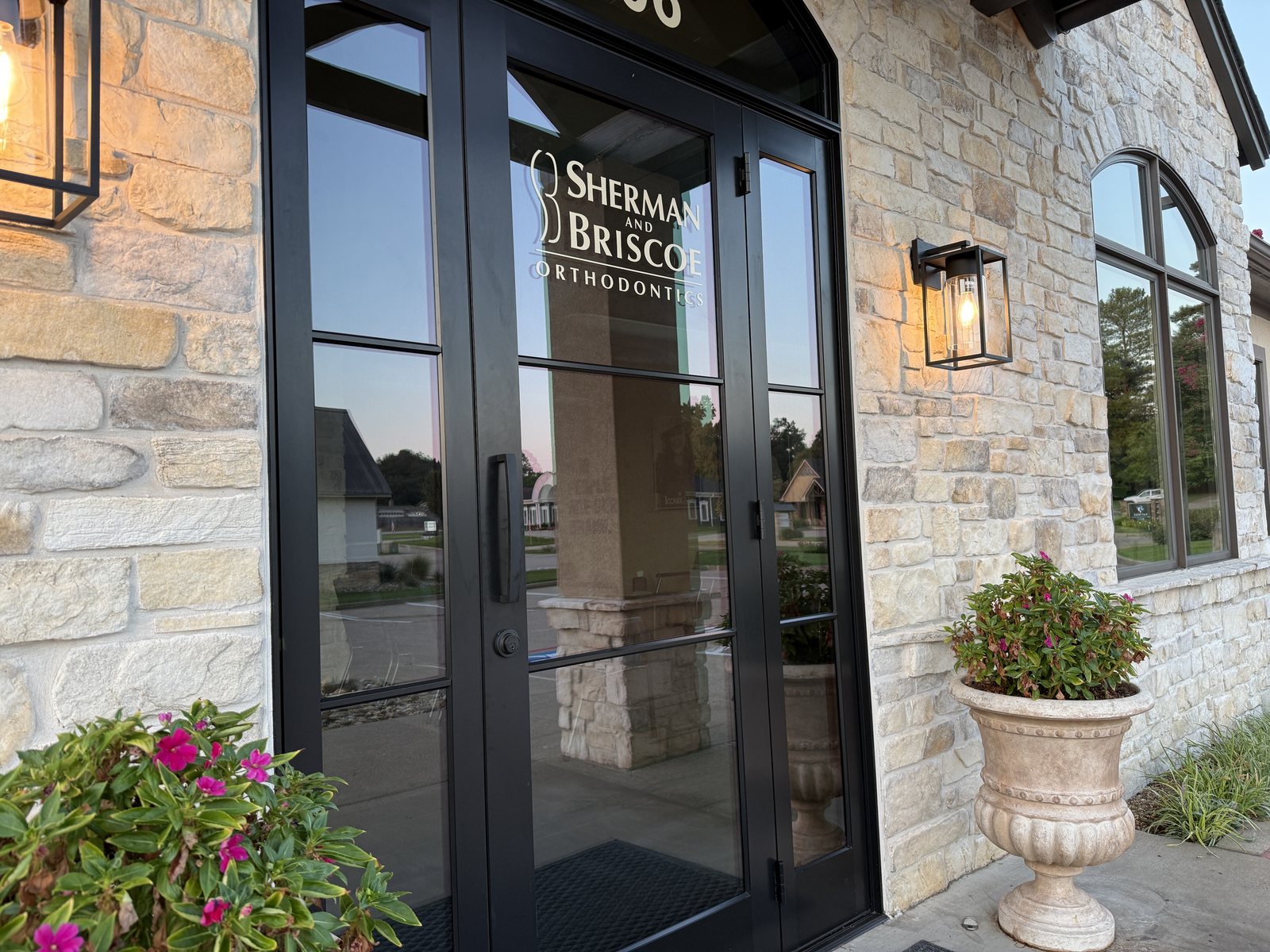 Glass entrance doors at the Longview office with the Sherman & Briscoe logo, warm lanterns, stone walls, and flowering planters.