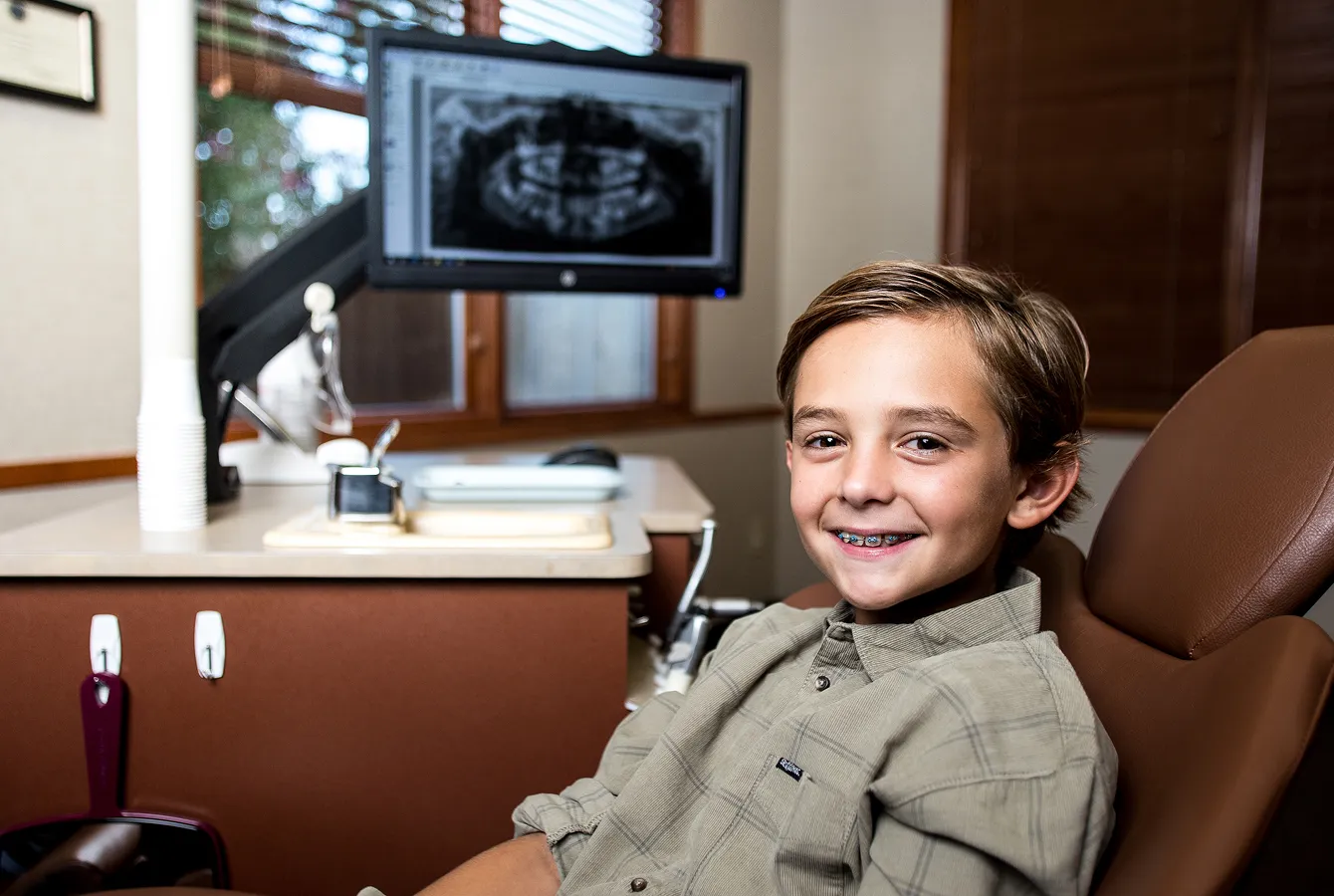 Young patient with braces smiling in an orthodontic treatment chair beside a panoramic dental X-ray.